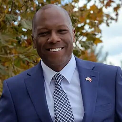 David Williams smiling outdoors in a blue suit and patterned tie with a flag pin, autumn foliage behind him.